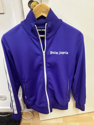 Palm Angels Track Jacket - Säljer en lila Palm Angels Track Jacket med vita dragkedjor och vita stripes längs ärmarna. Jackan har hög krage, två fickor med dragkedja och klassisk Palm Angels-logga på bröstet. Perfekt för dig som vill sticka ut med streetwear-vibe. Mycket bra skick, 9/10.