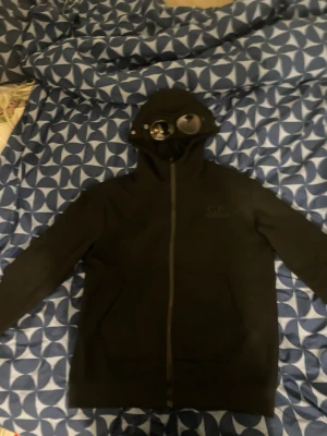 Svart hoodiejacka med goggles från C.P. Company - Svart hoodiejacka från C.P. Company med ikoniska goggles i huvan. Jackan har dragkedja framtill, två fickor och lång ärm. Materialet är mjukt och känns som bomull eller fleece. Perfekt för dig som vill sticka ut med streetwear-vibe.