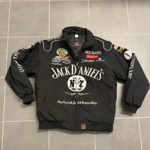 Jack Daniel's svart bomberjacka L - Svart bomberjacka från Jack Daniel's med stora broderade loggor och racing-inspirerade patches på bröstet och ärmarna. Jackan har dragkedja, ribbade muddar och krage, samt två fickor fram. Materialet är syntet och jackan har quiltad insida.