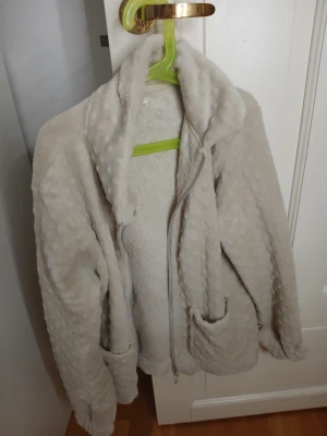 Beige teddyjacka från Pomp de Lux - Supermjuk och fluffig teddyjacka i beige från Pomp de Lux. Jackan har lång ärm, dragkedja framtill och två praktiska fickor. Materialet är fleece med en cool prickig struktur som ger extra mysfaktor. Perfekt för kalla dagar när du vill vara både chill och varm.