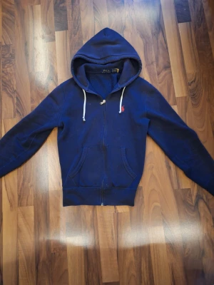 Marinblå hoodie från Polo Ralph Lauren XS - Snygg marinblå hoodie från Polo Ralph Lauren i storlek XS. Tröjan har huva med vita snören, dragkedja framtill och klassisk röd broderad logga på bröstet. Mjuk insida och två fickor framtill gör den både chill och praktisk. Snörena är lite sunkiga men allt annat är i bra skick. Du kan annars ta bort snörena eller köpa nya.
