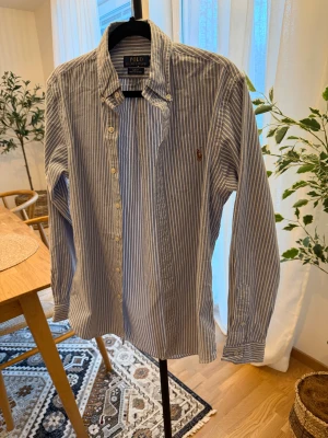 Blårandig skjorta Polo Ralph Lauren S - Slim fit stretch oxfordskjorta från Polo Ralph Lauren i blått och vitt med vertikala ränder. Klassisk krage, knappar framtill och broderad logga på bröstet. Långärmad och tillverkad i bomull med skön stretch. Perfekt för dig som gillar stilren och tidlös look.