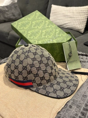 Gucci keps med GG-mönster och röd-blå detalj - Snygg beige och mörkblå keps från Gucci med klassiskt GG-mönster över hela kepsen. Har en röd och blå rand på sidan och justerbar rem baktill i svart skinn. Materialet är bomull och kepsen har en cool streetstil som sticker ut. Finns även i svart man kan få både denna och svart för 1300 kr 