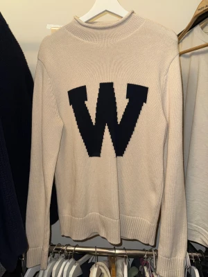 Beige stickad tröja med W från Whyred - Snygg beige stickad tröja från Whyred i 100% bomull. Framtill finns ett stort  W som ger en cool collegekänsla. Tröjan har långärmad passform, ribbade muddar och en lite högre hals. Perfekt för dig som gillar stilrena detaljer och vill vara stilren nu till vintern! Tröjan är som ny