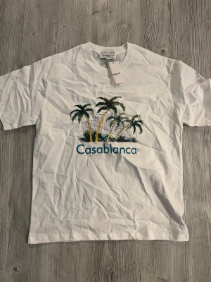Vit Casablanca t-shirt med palmtryck helt ny! - Vit t-shirt från Casablanca med färgglatt tryck av palmer och byggnad på bröstet samt tydlig logga. Klassisk rund hals och korta ärmar. Materialet är mjuk bomull som känns skönt mot huden. Perfekt för dig som gillar stilrena plagg med coola detaljer.