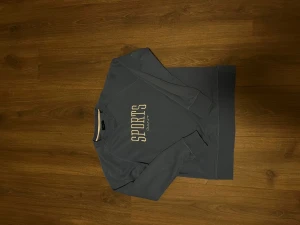 Blå sweatshirt från GANT med sytt tryck - Blå sweatshirt från GANT med stort broderat SPORTS-tryck i vitt på bröstet. Tröjan har rund hals, ribbade muddar och är tillverkad i mjuk bomull. Perfekt för dig som gillar sportig och clean stil.