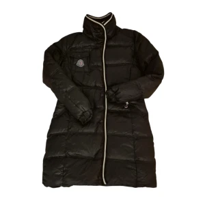 Svart dunjacka från Moncler women L - Säljer en svart dunjacka från Moncler med hög krage och vit kant längs dragkedjan. Jackan har quiltad design, långa ärmar och praktiska fickor med knappar och dragkedja. Moncler-logga på bröstfickan ger en exklusiv vibe. Perfekt för kalla dagar.