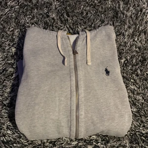Grå hoodie från Ralph Lauren - Säljer en stilren grå hoodie från Ralph Lauren med dragkedja och det klassiska logotypbroderiet på bröstet. Perfekt för en avslappnad look. En liten defekt är att löparen på kedjan har lossnat. Skick 7/10 st S 