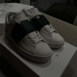 Valentino Garavani Open sneakers vit/svart - Snygga Valentino Garavani Open sneakers i vitt skinn med bred svart rem över mitten. Klassisk rund tå, platt sula och vita snören. Priset kan diskuteras,