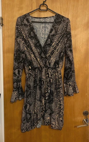 Beige och svart omlottklänning med volang - Säljer en kort omlottklänning med beige och svart leopardmönster. Klänningen har långa ärmar med volang vid ärmslut och nederkant, samt knyt i midjan för snygg passform. Materialet är lätt och luftigt. Ny. Stär inte storlek pà men när jag köpte den var den M. :)