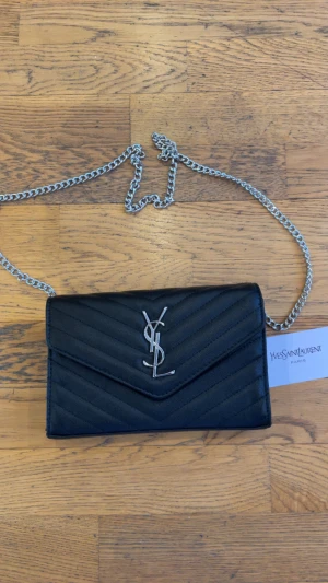 Svart quiltad axelväska från Saint Laurent - Snygg svart axelväska från Saint Laurent med silverfärgad kedja och ikoniskt YSL-emblem framtill. Väskan är quiltad i ett chevronmönster och har flera fack inuti, inklusive ett rött innerfack med dragkedja. Perfekt storlek för det viktigaste.