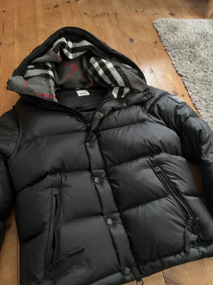 Svart pufferjacka från Burberry - Säljer en svart pufferjacka från Burberry med huva och klassiskt rutigt foder i grått, vitt, svart och rött. Jackan har dragkedja och knappar framtill, två snedställda fickor med dragkedja och en patch med Burberry London England på ärmen. Perfekt för kalla dagar.