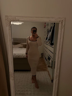 Beige ribbad långklänning med svarta detaljer - Säljer en beige ribbad långklänning med lång ärm och tight passform. Klänningen har fyrkantig ringning och svarta vågiga detaljer vid halsringning och nederkant. Perfekt för dig som gillar stilrena och trendiga plagg.