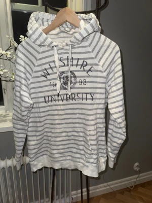 Randig hoodie från L.O.G.G. H&M - Snygg vit och grårandig hoodie från L.O.G.G. H&M med tryck 'Wilshire University 1993' på bröstet. Tröjan har huva med snörning, känguruficka och långa ärmar. Perfekt för chill dagar och har en klassisk college-vibe.