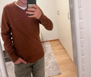 Brun tröja  Tommy Hilfiger - Snygg brun v-ringad tröja från Tommy Hilfiger med diskret logga på bröstet. Tröjan är långärmad och har ribbade muddar vid ärmslut och nederkant. Tillverkad i mjuk bomull, perfekt för dig som gillar klassisk stil med en modern touch. Den är bara använd en gång och är 92% bomull och 8% chasmire. Skriv ifall ni har frågor. 
