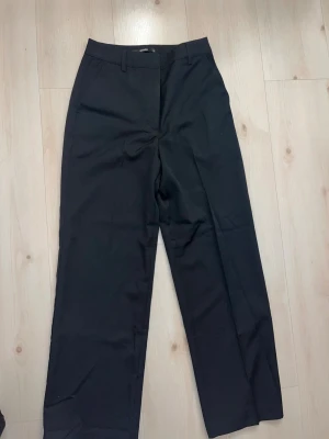 Svarta kostymbyxor från Bik Bok - Svarta kostymbyxor från Bik Bok med klassisk rak passform och midwaist. Byxorna har bälteshällor, sidfickor och är tillverkade i ett slätt material som ger en clean look. Perfekta för dig som gillar stilrena och enkla plagg.