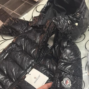 Svart pufferjacka från Moncler barn 1-3 år  - Säljer en svart glansig pufferjacka från Moncler med huva och dragkedja. Jackan har Moncler-logga på ärmen och flera praktiska fickor med dragkedja. Perfekt för kalla dagar när du vill hålla stilen.