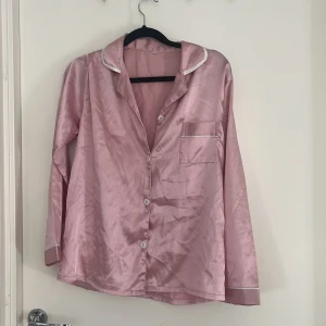 Rosa satinpyjamas med vita detaljer - Snygg rosa pyjamasskjorta i satin med vita kantdetaljer och knappar framtill. Skjortan har klassisk krage, bröstficka och långa ärmar. Matchande byxor med resår i midjan ingår. 