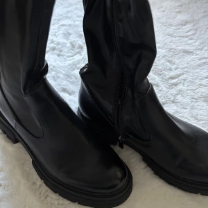 Svarta höga boots med dragkedja från  - Säljer ett par svarta höga boots med chunky sula och dragkedja på insidan. Använda 1-2 gånger, som nya! Sitter smickrande på benet, passar mig med större vader. Storlek 38 från marco tozzi. Stilrena och lätta att matcha med det mesta. Följsamma längs benet. Slutar under knät. Nypris 1800kr.