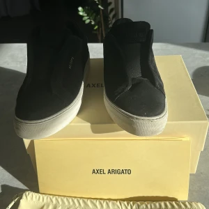 Svarta sneakers från Axel Arigato - Säljer ett par svarta slip-on sneakers från Axel Arigato med vit platt sula. Skorna har minimalistisk design, rund tå och är tillverkade i mocka med elastiska paneler för enkel påtagning. Perfekta för dig som gillar stilrena och bekväma skor.Box,påse och klistermärken ingår