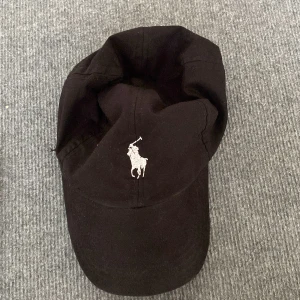Svart keps från Polo Ralph Lauren - Svart keps från Polo Ralph Lauren med klassisk vit broderad logga framtill och 'POLO' broderat baktill. Justerbar passform med kardborreband. Tillverkad i bomull, perfekt för en clean och sportig look.