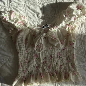 Blommig volangblus från Gina Tricot - Supersöt blommig blus från Gina Tricot i vitt med rosa och gröna blommor. Toppen har volangdetaljer vid ärmar och nederkant, smockad midja och knyt framtill. Perfekt för en romantisk look med sina puffärmar och lätta material.