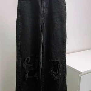 Svarta wide jeans med revor från Bershka - Svarta jeans från Bershka, modell THE 90's, med breda ben och stora slitningar på knäna. Jeansen har klassisk femficksdesign och är tillverkade i denim. Perfekta för dig som gillar en avslappnad och trendig look.