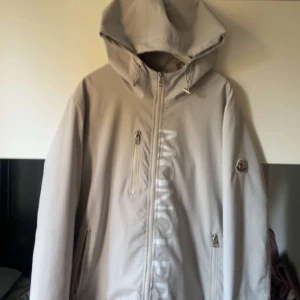 Beige vindjacka från Moncler med huva - Snygg beige vindjacka från Moncler med stor huva och dragkedja framtill. Jackan har Moncler-logga på ärmen, diskret bröstficka med dragkedja och två sidofickor. Stort Moncler-tryck längs dragkedjan ger en clean och exklusiv vibe.