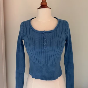 Blå ribbad långärmad topp - Säljer en blå ribbad topp med lång ärm och croppad passform. Toppen har rund halsringning och knappar framtill som ger en snygg detalj. Perfekt för dig som gillar en enkel men stilren look.