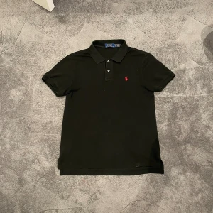 Svart pikétröja Polo Ralph Lauren XL - Svart pikétröja från Polo Ralph Lauren i slim fit-modell. Klassisk krage, korta ärmar och knäppning framtill. Ikoniska röda loggan broderad på bröstet. Perfekt för en clean och stilren look.     Axel-43 Längd- 67