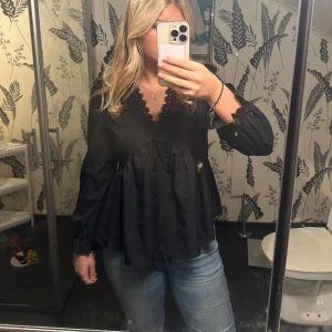 Svart blus med spets och v-ringning - Svart blus med v-ringning och fina spetsdetaljer längs urringningen. Blusen har lång ärm med lätt puff och resår vid ärmslut. Modellen är lös och har en volangliknande nederdel som ger en flowy känsla. Perfekt att styla med jeans eller kjol.
