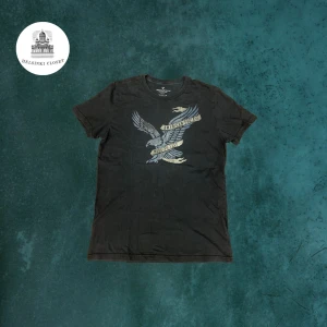 Svart t-shirt från American Eagle - Svart t-shirt från American Eagle Outfitters med coolt tryck av en örn och texten 'American Free'. Klassisk passform och rund halsringning. Perfekt för dig som gillar streetstyle och vill ha en bekväm och snygg tee.