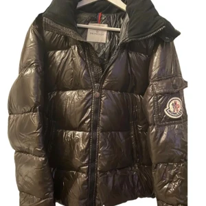Svart pufferjacka från Moncler - Snygg svart pufferjacka från Moncler med glansig finish och stor huva. Jackan har Moncler-logga på ärmen, dragkedja framtill och quiltad design. Perfekt för kalla dagar och ger en riktigt trendig look. Inköp från vestiaire, därför ingen kvitto! 