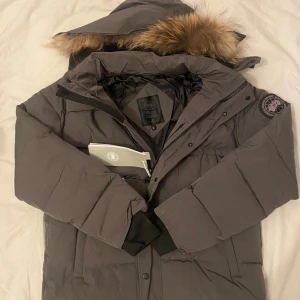Grå Canada Goose black badge  - Säljer en grå dunjacka från Canada Goose med huva och snygg pälsdetalj. Jackan har svarta muddar, dragkedja och knappar framtill samt logotyp på ärmen. Insidan är fodrad i svart och jackan har en loose passform. Perfekt för kalla vinterdagar.