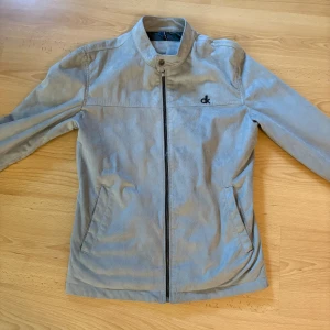 Beige mockajacka Calvin Klein Jeans - Snygg grå mockajacka från Calvin Klein Jeans med stilren ståkrage och svart CK-logo på bröstet. Jackan har dragkedja framtill, två fickor och är helfodrad. Perfekt för dig som gillar minimalistisk design och mjuka material.