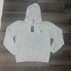 Ralph Lauren hoodie - helt ny! Snygg ljusgrå hoodie från Polo Ralph Lauren med klassisk broderad logga på bröstet. Tröjan har huva med snörning, känguruficka och ribbade muddar. Tillverkad i mjuk bomullsmix som är perfekt för höst och vinter. ÄKTA! Axellbredd 53cm, längd 63cm!