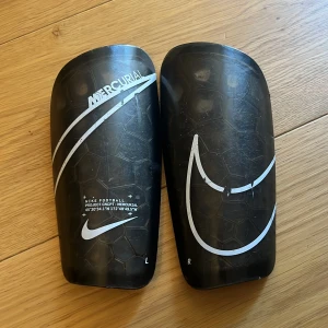 Nike Mercurial benskydd svart - Säljer ett par svarta Nike Mercurial benskydd med vit logga och coolt hexagonmönster. De är formade för att sitta skönt på benet och har en slitstark yta med snygga detaljer. Perfekt för dig som vill ha stil och skydd på fotbollsplanen.