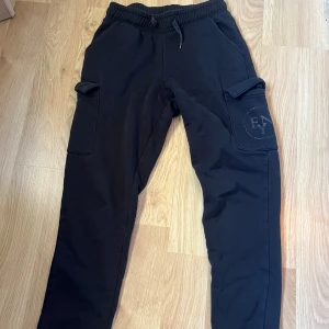 Svarta cargobyxor från Ellen New York - Svarta cargobyxor från Ellen New York med elastisk midja och snörning. Byxorna har stora fickor på sidorna och en diskret logga på ena benet. Tillverkade i mjukt material som är skönt att ha på sig hela dagen.