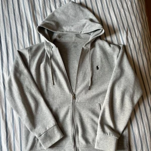 Grå hoodie från Polo Ralph Lauren - Snygg grå hoodie från Polo Ralph Lauren med dragkedja framtill och klassisk huva med snörning. Tröjan har långa ärmar, två fickor och den ikoniska svarta loggan broderad på bröstet. Perfekt för en chill och stilren look.