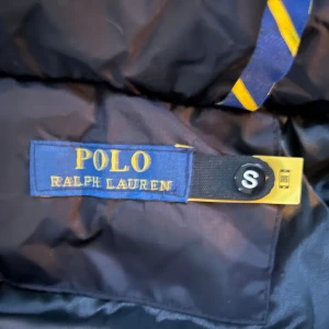 Svart pufferjacka från Polo Ralph Lauren - Svart pufferjacka från Polo Ralph Lauren med huva och röda loggan broderad på bröstet. Jackan har flera dragkedjeförsedda fickor och quiltad design. Perfekt för kalla dagar och riktigt snygg streetstil.