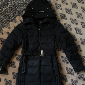 Svart dunjacka från Canada Snow - Snygg svart dunjacka från Canada Snow med huva, dragkedja och markerad midja med elastiskt bälte. Jackan har quiltad design, långa ärmar och praktiska fickor med dragkedja. Perfekt för kalla dagar och riktigt stilren look.