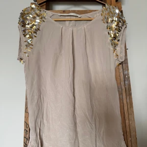 Beige blus med paljetter från Custommade - Beige blush side från Custommade med korta ärmar och guld- och silverpaljetter på axlarna. Storlek S