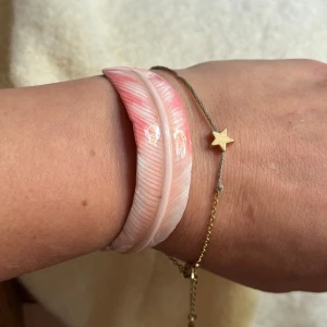 Rosa fjäderformad armband / Banglet - Armband i form av en fjäder, med rosa och vita nyanser. Armbandet har en metallbåge i guldton och fjädern är glansig med detaljerad struktur. Storleken går att reglera. 