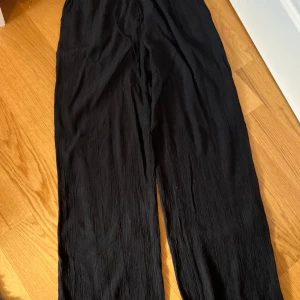 Svarta vida byxor i crinkle-tyg - Säljer ett par svarta byxor med vid passform och snyggt crinkle-material. Byxorna har rak siluett och är perfekta för en chill och trendig look. Passar dig som gillar bekväma och stilrena plagg.