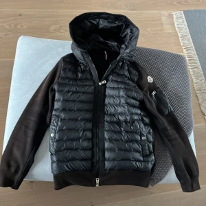 Svart pufferjacka från Moncler med huva - Snygg svart pufferjacka från Moncler med quiltad framsida och huva. Jackan har släta ärmar, dragkedja framtill och en liten ficka med dragkedja på ena ärmen. Perfekt för dig som vill ha en stilren och sportig look.