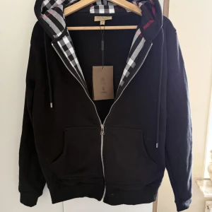 Burberry kofta - Lyxig hoodie från Burberry i svart med den klassiska rutiga huvan som detalj. Ikonisk och tidlös design med högsta kvalitet – perfekt för både vardag och mer dressade tillfällen.    Helt ny – med originaltag kvar   Exklusiv Burberry-detalj i huvan     Snabb leverans & trygg affär
