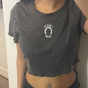 Svart croppad t-shirt med tryck - Säljer en svart croppad t-shirt med vit text och hästsko-tryck på bröstet där det står 'Just in luck'. T-shirten har korta ärmar, rund hals och rå kant nedtill för en chill vibe. Perfekt för dig som gillar avslappnad stil.