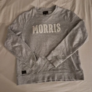 Grå sweatshirt från Morris i bomull - Snygg grå sweatshirt från Morris med stora, fluffiga bokstäver framtill. Tröjan har rund halsringning, ribbade muddar och är tillverkad i mjuk bomull. Perfekt för en chill och stilren look.