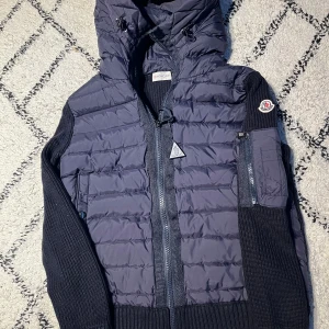 Mörkblå Moncler dunjacka med huva - Snygg mörkblå dunjacka från Moncler med stickade ärmar och quiltad front. Jackan har huva med dragsko, dragkedja framtill och Moncler-logga på ärmen. Perfekt för kyliga dagar och har en modern, sportig look. Pris kan diskuteras vid snabb affär!