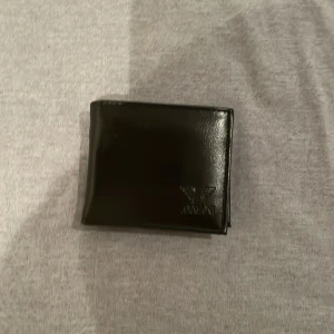 Armani plånbok i svart läder - Elegant svart läderplånbok från Armani med flera kortfack, myntficka och fönster för ID. Plånboken är i fint skick utan synligt slitage och passar perfekt för dig som söker en stilren och praktisk accessoar.